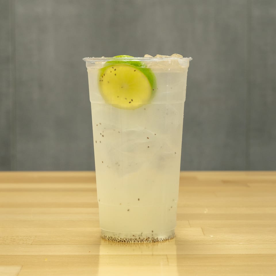 Chilled pale fruit drink with lime slices and basil seeds in a clear cup on a wooden counter.