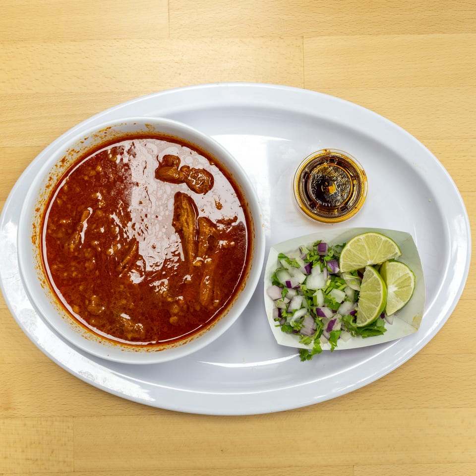 Bowl of red stew served with chopped herbs, onions, lime wedges, and dipping sauce on a white tray.