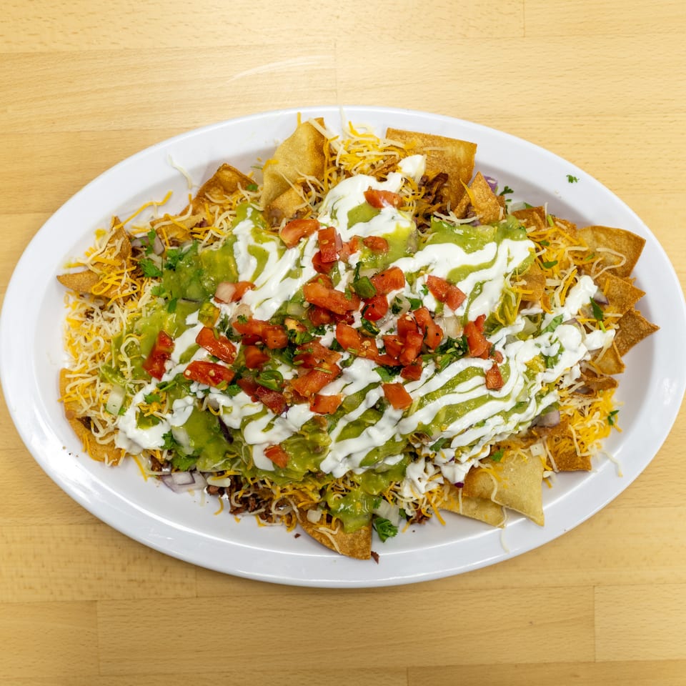 Loaded nachos topped with melted cheese, guacamole, sour cream, diced tomatoes, onions, and cilantro on a white platter.