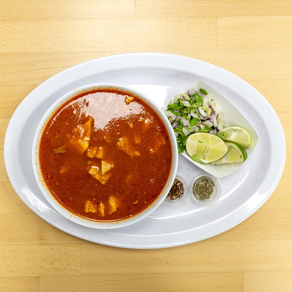 Bowl of rich red soup served with chopped onions, cilantro, lime wedges, and small condiment cups on a white platter.
