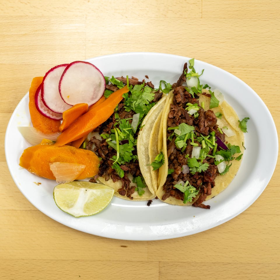 Plate of soft corn tacos filled with seasoned meat, cilantro, and onions, served with pickled carrots, radishes, and lime wedges.
