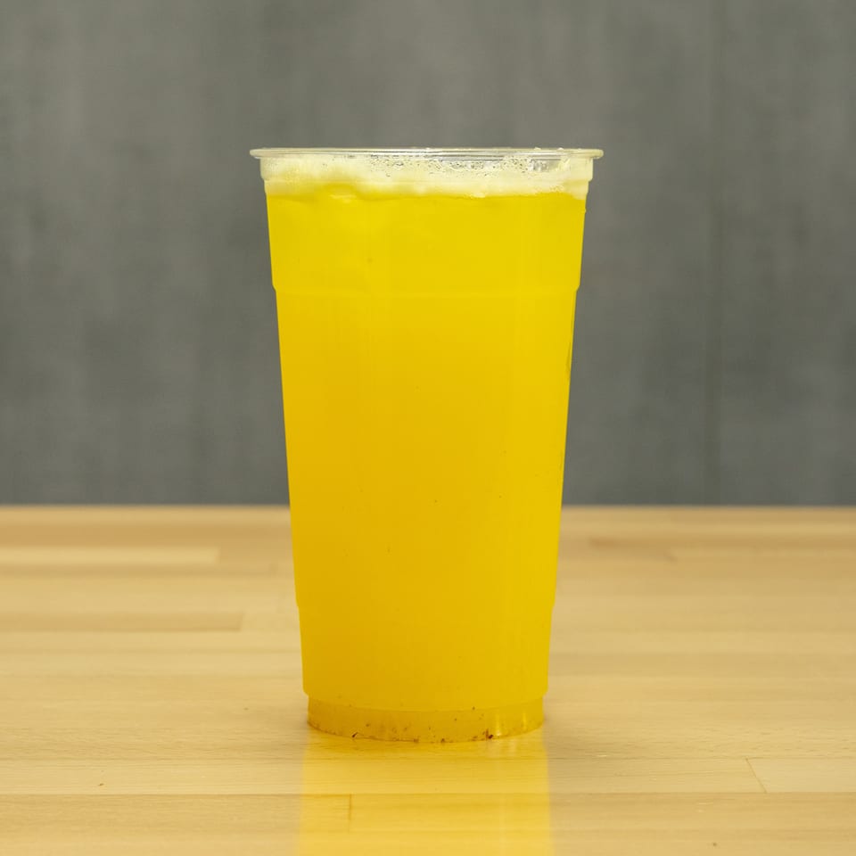 Bright yellow beverage in a clear plastic cup with frothy foam, standing on a wooden table against a gray background.