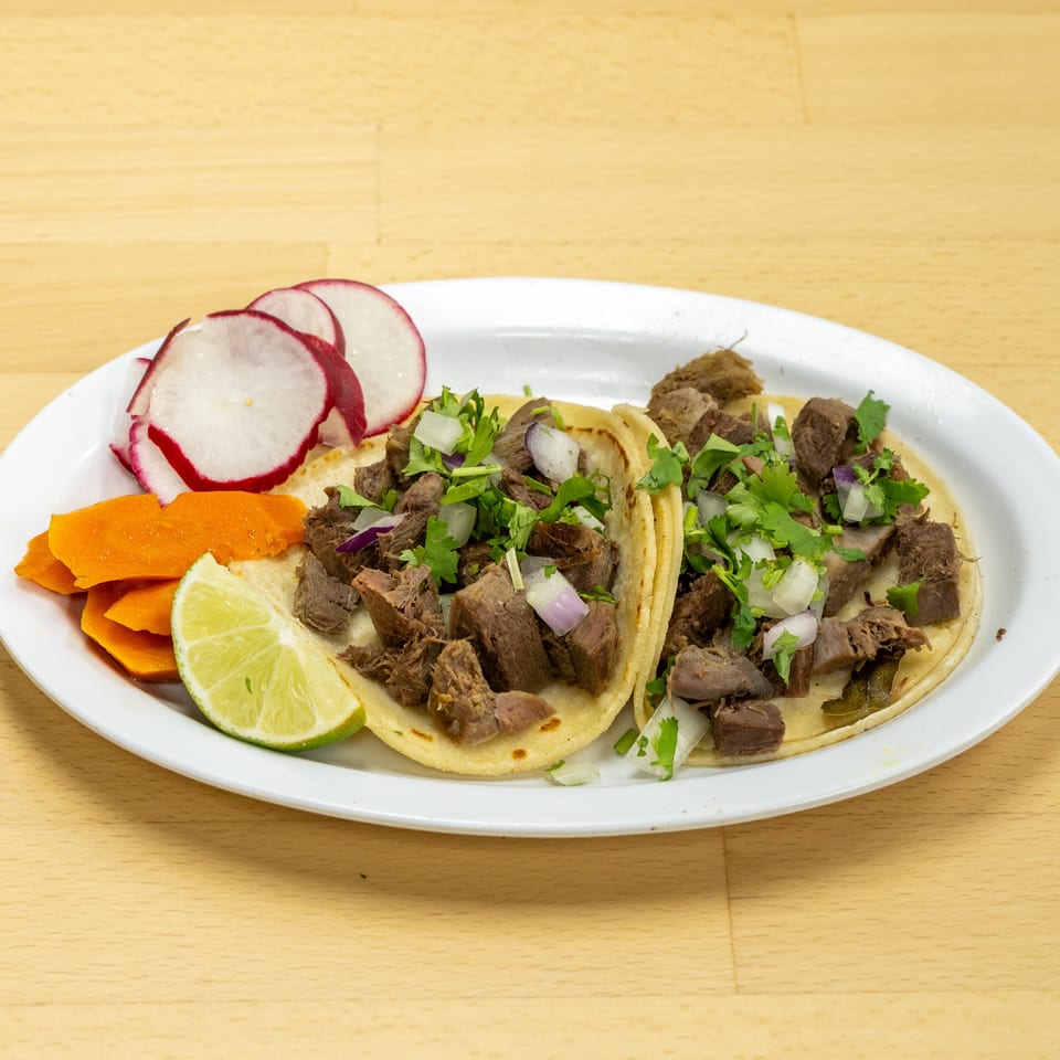 Plate of beef tacos topped with cilantro and onions, served with lime, radishes, and carrots.