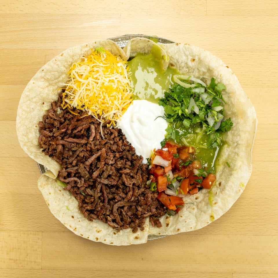 Overhead view of a loaded flour tortilla with seasoned beef, shredded cheese, guacamole, sour cream, pico de gallo, cilantro, and onions.