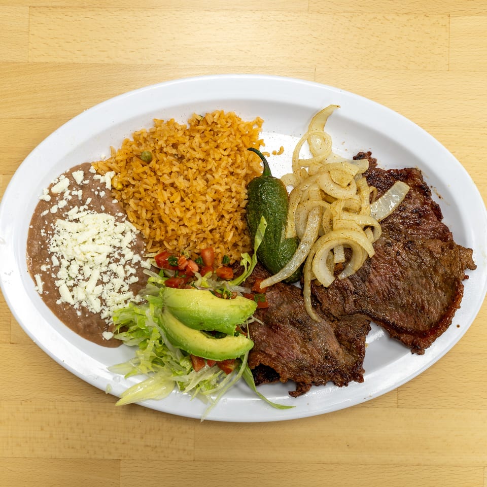 Grilled steak plate with Mexican rice, refried beans topped with cheese, avocado salad, grilled onions, and a roasted pepper.