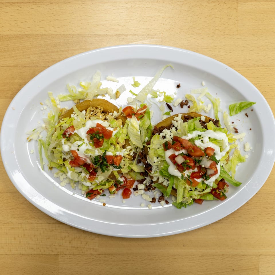 Two loaded tacos topped with lettuce, ground beef, diced tomatoes, cheese, and white sauce on a large white plate.