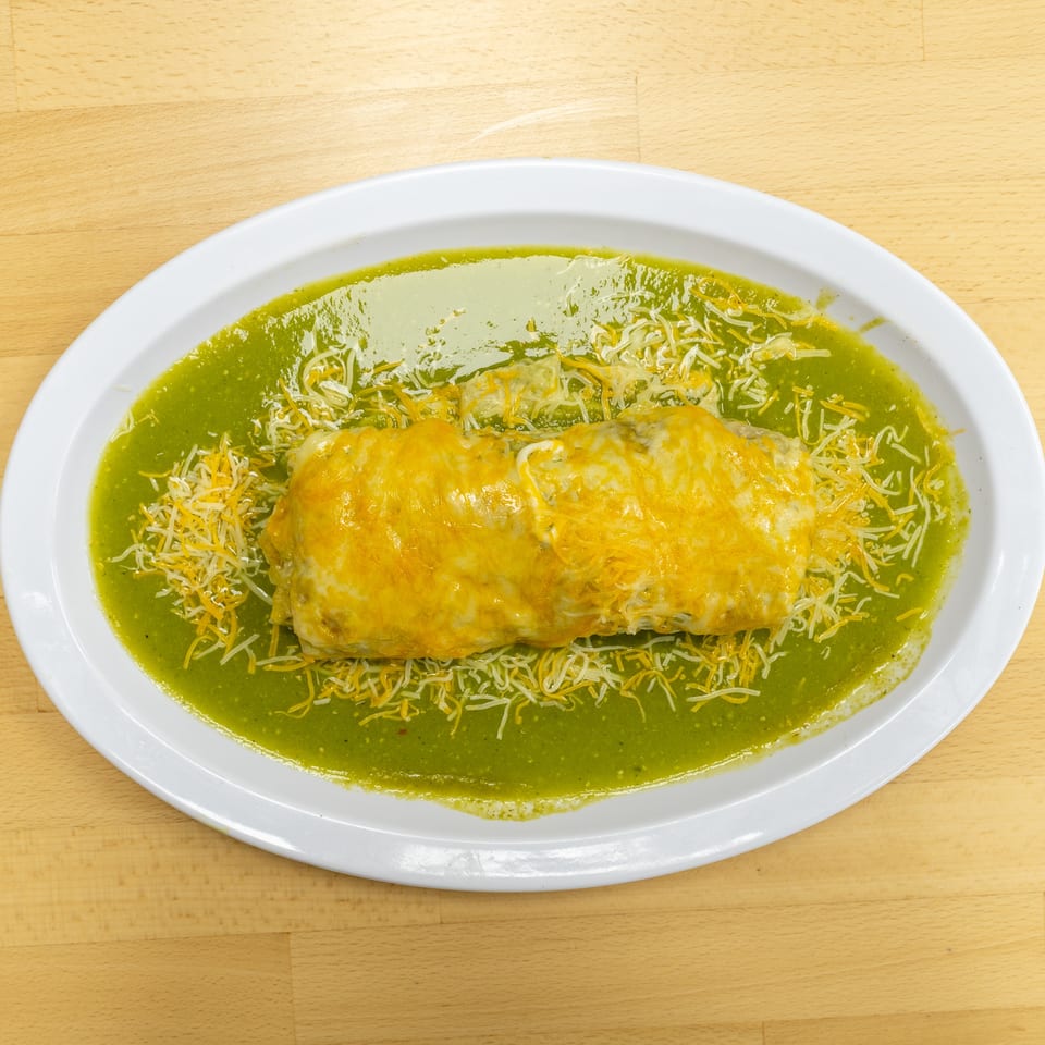 Cheesy enchiladas surrounded by shredded cheese and green sauce on a white oval plate atop a wooden table.