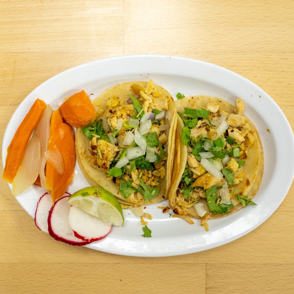 Two chicken tacos topped with cilantro and onions, served with pickled carrots, onions, radishes, and lime on a white plate.