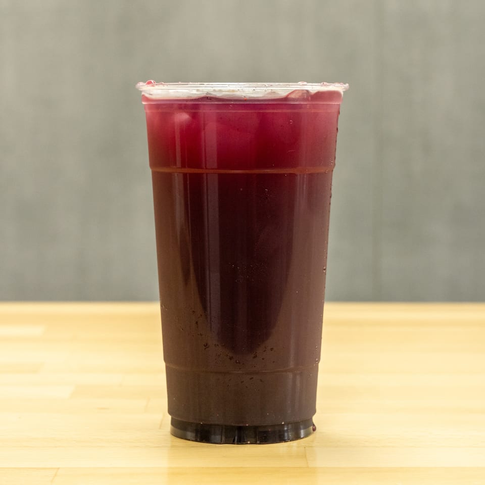 Tall clear plastic cup filled with dark red iced beverage on a light wooden table.