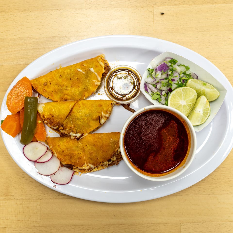 Plate of golden birria tacos with consommé, chopped onion and cilantro, lime wedges, pickled vegetables, and radishes.