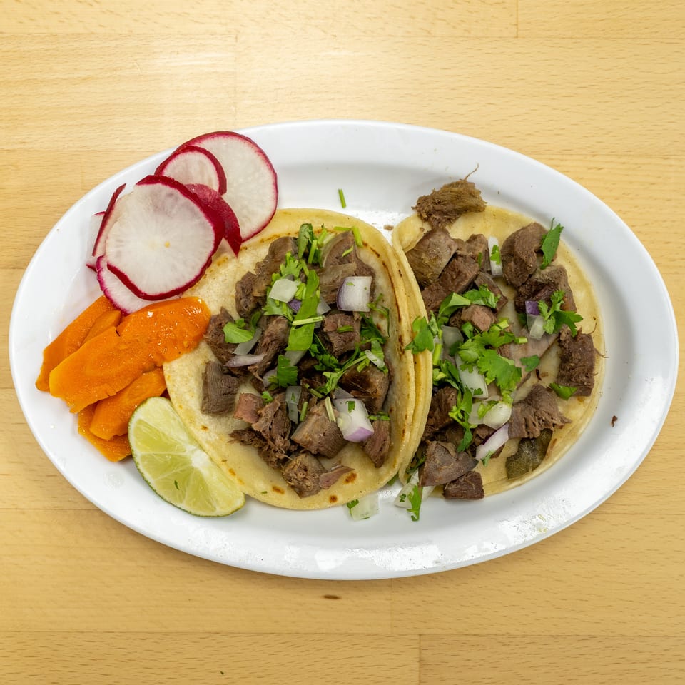 Two soft tacos filled with chopped meat, onions, and cilantro, served with radishes, carrots, and lime.