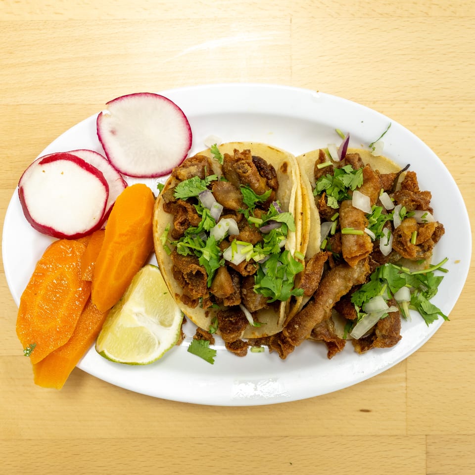 Plate of crispy meat tacos topped with cilantro and onions, served with radishes, carrots, and lime wedges.